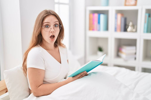 Νεαρή καυκάσια γυναίκα κάθεται στο κρεβάτι στο σπίτι διαβάζοντας ένα βιβλίο φοβισμένη και έκπληκτη με ανοιχτό το στόμα για έκπληξη, δυσπιστία πρόσωπο  - Φωτογραφία, εικόνα