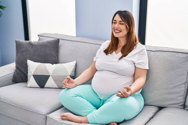 Young pregnant woman doing yoga exercise sitting on sofa at home - Valokuva, kuva