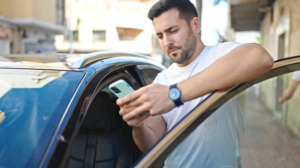 Young hispanic man using smartphone leaning on car at street - Foto, imagen
