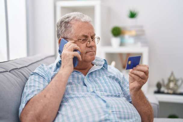 Middle age grey-haired man talking on smartphone holding credit card sitting on sofa at home - Photo, Image