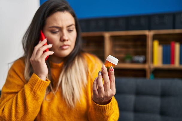 Young beautiful plus size woman talking on smartphone holding pills at home - Fotoğraf, Görsel