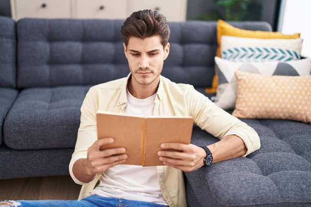 Fiatal spanyol férfi könyvet olvas, otthon ül a padlón. - Fotó, kép