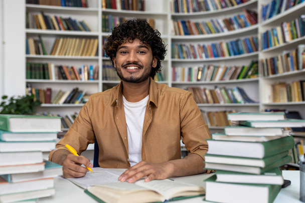 Happy and smiling student studying inside academic library, man smiling and looking at camera doing homework among books. - Zdjęcie, obraz