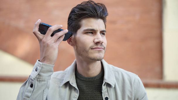 Young hispanic man listening audio message by the smartphone with serious expression at street - Фото, изображение