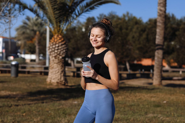 Active young fit woman in wireless headphones drinks water from bottle after exercising outdoors at palm trees park. Happy girl in sportswear quenches thirst and cools down after running or workout. - Фото, зображення