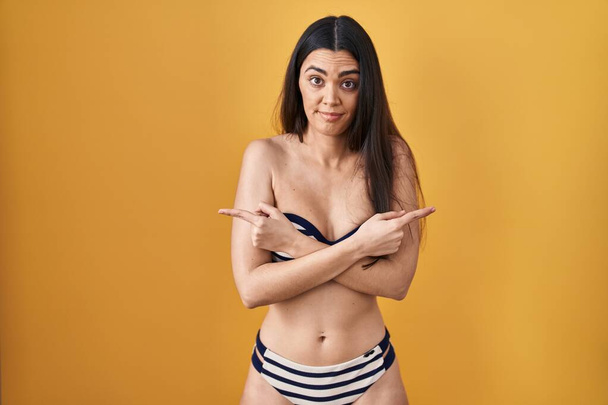 jonge brunette vrouw dragen bikini over gele achtergrond wijzend naar beide zijden met vingers, verschillende richting oneens  - Foto, afbeelding