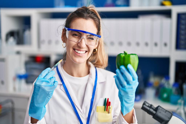 Mujer hispana joven trabajando en laboratorio científico con verduras guiñando el ojo mirando a la cámara con expresión sexy, cara alegre y feliz.  - Foto, Imagen