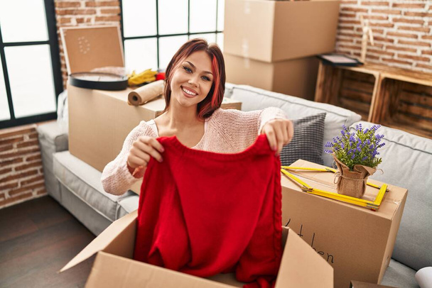 Mujer joven caucásica sonriendo confiado desembalaje ropa de cartón caja en nuevo hogar - Foto, imagen