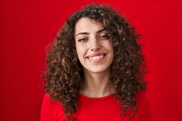 Hispanic woman with curly hair standing over red background smiling with a happy and cool smile on face. showing teeth.  - Zdjęcie, obraz