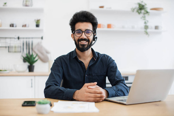 Portrait of smiling person wearing headset with microphone posing with documents and portable computer. Cheerful arabian consultant offering guidance to clients over digital platform from home. - Φωτογραφία, εικόνα