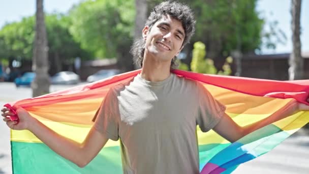 Jonge Spaanse man glimlacht vol vertrouwen met regenboogvlag op straat - Video
