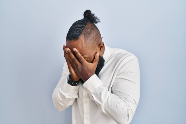 Afrikanischer Mann, der vor blauem Hintergrund steht, mit traurigem Gesichtsausdruck, der das Gesicht mit Händen bedeckt, während er weint. Depressionskonzept.  - Foto, Bild