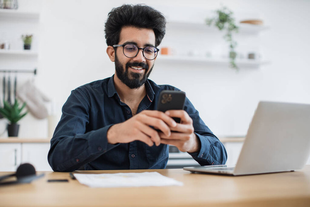 Positive indian male in spectacles holding smartphone while staying in modern workplace at home. Efficient freelance worker texting message to colleague while performing project for employer indoors. - Fotografie, Obrázek