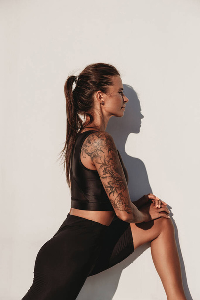 Beautiful young fit tanned tattooed caucasian woman warming up on the white wall background on summer sunny day - Zdjęcie, obraz