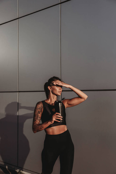 Beautiful young fit tanned caucasian woman with tattoo holding a protein shaker or water bottle on grey wall background on sunset - Zdjęcie, obraz
