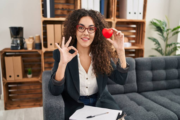 Junge hispanische Frau, die in der Beratungsstelle an Paartherapie arbeitet, macht Okay-Zeichen mit den Fingern, lächelt freundlich und gestikuliert ein hervorragendes Symbol  - Foto, Bild