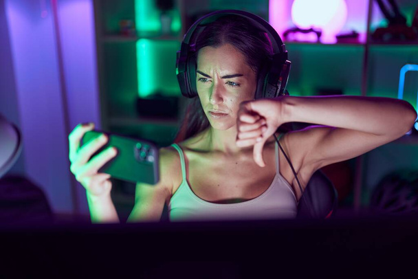 Junge brünette Frau spielt Videospiele mit Smartphone mit wütendem Gesicht, negatives Zeichen für Abneigung mit gesenktem Daumen, Ablehnungskonzept  - Foto, Bild