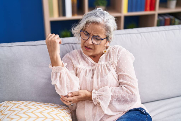 Grauhaarige Frau mittleren Alters sitzt auf Sofa und leidet zu Hause unter Ellbogenschmerzen - Foto, Bild