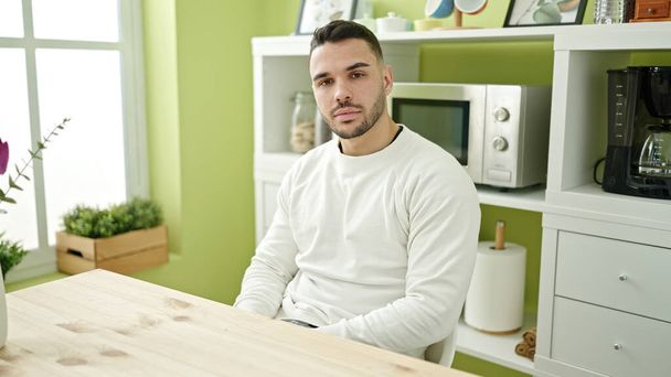 Jeune homme hispanique assis sur la table avec une expression sérieuse à la salle à manger - Photo, image