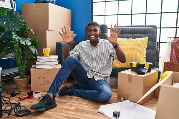 アフリカ系アメリカ人の男が新しい家で床に座って指の番号10を示すと指摘しながら、自信と幸せ笑顔.  - 写真・画像