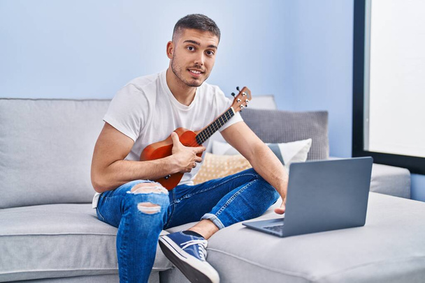 Joven hombre hispano que tiene clase de ukelele en línea sentado en el sofá en casa - Foto, imagen