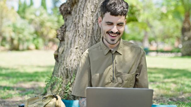 Young hispanic man using laptop sitting on floor smiling at park - Photo, Image