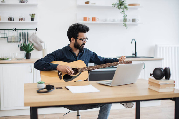 Young male musician in dark shirt tuning acoustic guitar using laptop on writing desk in modern apartment. Focused indian male practising audio exercises on string instrument via online tutorial. - Photo, Image