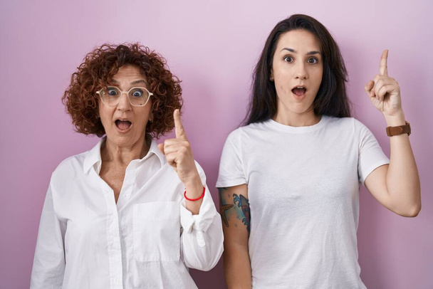 İspanyol anne ve kızı pembe arka plan üzerine gündelik beyaz tişört giyip başarılı bir fikirle işaret ediyorlar. Heyecanlı ve mutlu. Bir numara..  - Fotoğraf, Görsel