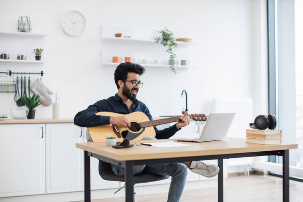 Talented indian musician holding acoustic guitar while analyzing information on laptop screen in bright dining room. Young bearded composer writing new song using digital devices and instrument. - Foto, Imagen