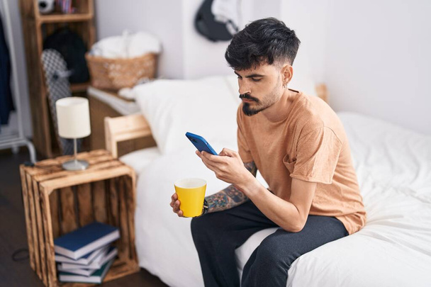 Young hispanic man using smartphone drinking coffee at bedroom - Photo, Image