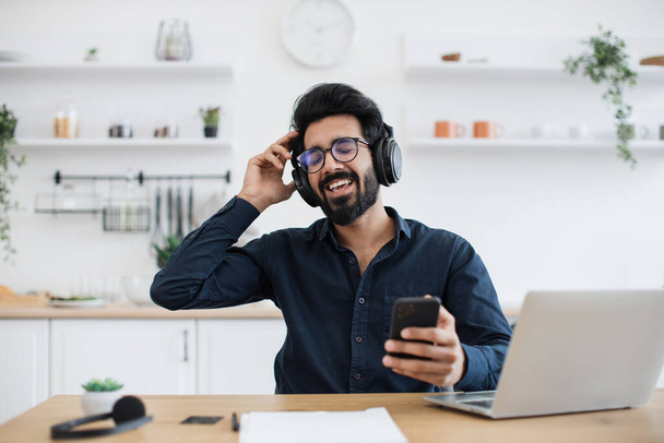 Easygoing arabian person in spectacles showing great enthusiasm for audio delivered through wireless devices indoors. True music lover enjoying sound quality of pure performance in headphones. - Fotó, kép