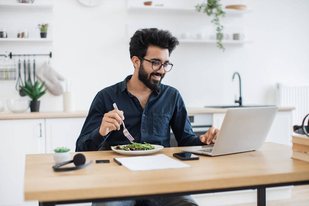 Smiling mature man in business casual clothes eating healthy salad while looking at laptop screen in kitchen interior. Indian remote employee having lunch break while doing home-based job at midday. - Foto, imagen