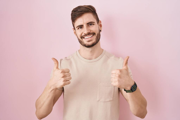Латиноамериканець з бородою, що стоїть над рожевим фоновим знаком успіху, робить позитивний жест рукою, великі пальці посміхаються і щасливі. Веселий вираз і жест переможця..  - Фото, зображення