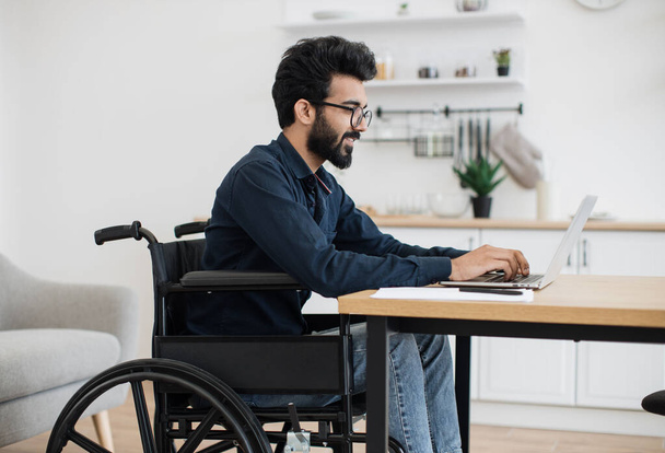 Close up view of smiling wheelchair user typing on portable computer while staying in distant workplace. Cheerful professional performing business project while doing full-time job from home. - Foto, immagini