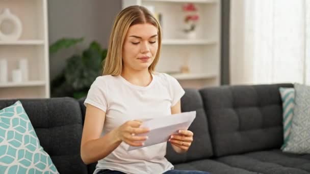 Giovane donna bionda lettera di lettura seduta sul divano con espressione preoccupata a casa - Filmati, video