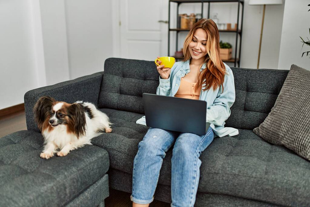 Молода біла жінка використовує ноутбук п'є каву, сидячи на дивані з собакою вдома
 - Фото, зображення