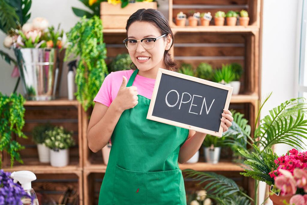 Hispanic young woman working at florist holding open sign smiling happy and positive, thumb up doing excellent and approval sign  - Photo, Image