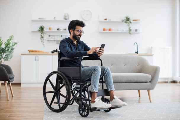 Full length view of indian male with disability using smartphone while resting in modern kitchen of apartment. Joyful person in glasses and casual clothes texting message on digital device at home. - Foto, immagini