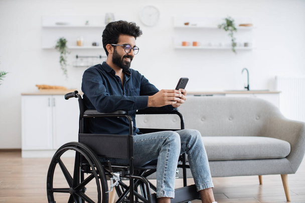 Smiling arabian adult in wheelchair looking at cell phone screen while having rest in comfortable dining room. Cheerful positive man checking social media using internet connection in apartment. - Valokuva, kuva