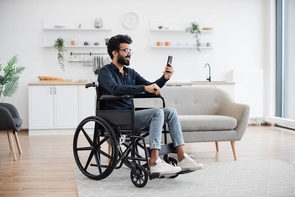 Cheerful wheelchair user in spectacles and casual outfit utilizing phone webcam while enjoying free time indoors. Bearded indian male receiving online conference call via mobile application about job. - Foto, imagen