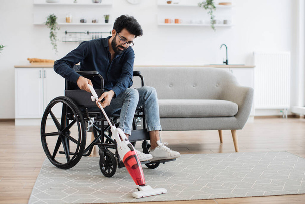 Helpful young man with disability holding cordless vacuum cleaner while sitting in wheelchair in open-plan kitchen. Active person in casual outfit collecting dust and dirt from carpet in apartment. - Fotografie, Obrázek