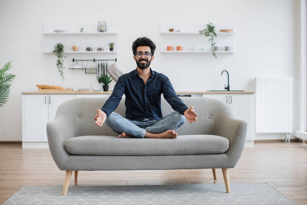 Fiatal mosolygós személy alkalmi ruhák ül keresztbe lábú kanapén modern apartman nyitott terű konyhával. Relaxált indiai férfi meditál ujjaival gyan mudra a napsütéses napon otthon. - Fotó, kép