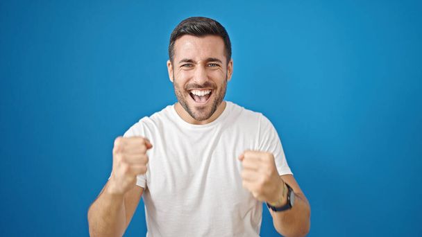 Молодий іспаномовний чоловік посміхається впевнено з виразом переможця над ізольованим синім тлом
 - Фото, зображення
