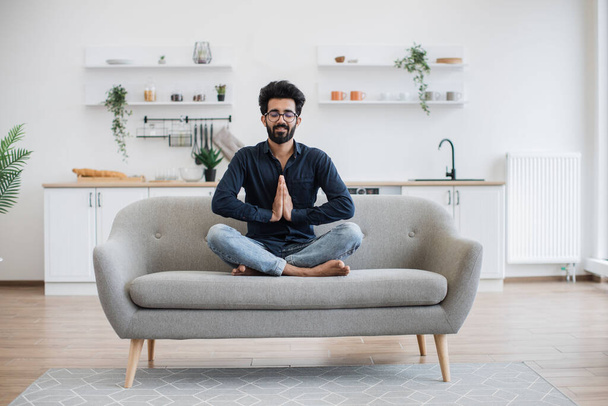 Young bearded man in jeans and dark shirt holding hands in anjali mudra while stretching in yoga pose on couch. Attractive indian adult relieving stress while sitting cross-legged in home interior. - Φωτογραφία, εικόνα