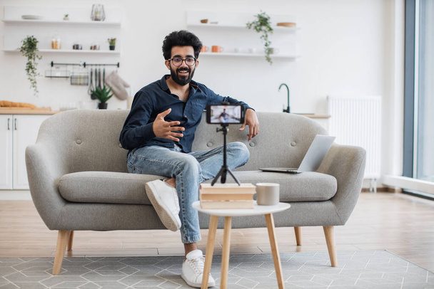 Front view of cheerful content maker in casual wear recording video to followers on modern smartphone. Young indian man in glasses with laptop sitting on comfortable sofa in living room. - Foto, Bild