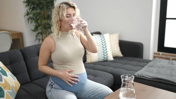 Joven embarazada bebiendo vaso de agua sentada en el sofá en casa - Foto, imagen