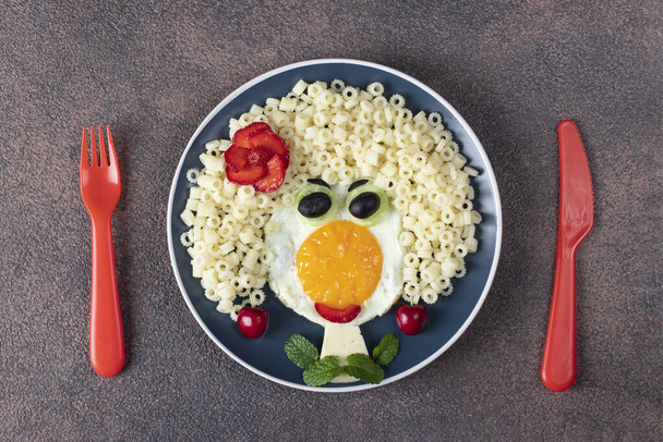 Fun Food voor kinderen - lachend gezicht meisje van gebakken eieren, pasta ditalini, olijven, aardbei en komkommer op blauwe plaat - Foto, afbeelding