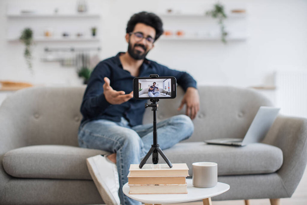 ラップトップでソファに座っている間、現代のスマートフォンで黒いひげの録音ビデオと正のインドの影響力。幸せな若い男はオンライン加入者と彼の成功を共有する. - 写真・画像