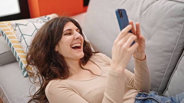 Junge schöne hispanische Frau mit Smartphone zu Hause auf Sofa liegend - Foto, Bild