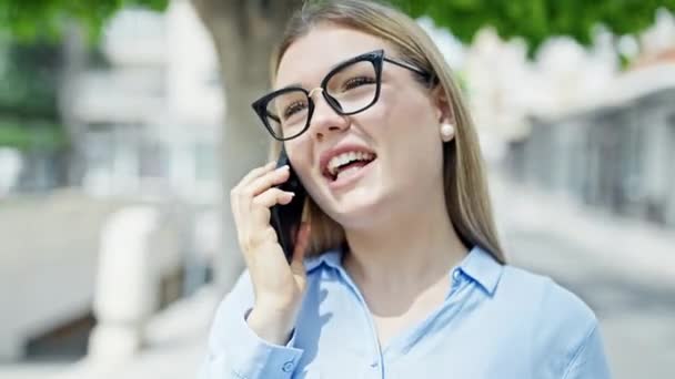 Nuori blondi nainen liiketoiminnan työntekijä hymyilee luottavainen puhuu älypuhelimeen puistossa - Materiaali, video